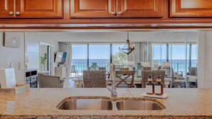 A kitchen with granite countertops, stainless steel sink, wooden cabinets, and a view of the ocean through large windows.