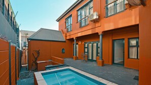 A swimming pool area with a tiled deck, a small seating area, and a building with orange walls and multiple windows. - Albatross Guest house (Cape Town)