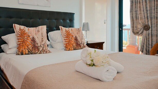 A neatly made bed with floral pillows, a towel, and a lamp beside it. - Albatross Guest house (Cape Town)