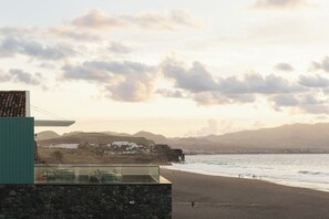 A coastal building with a stone facade, a balcony with chairs, and a view of the beach and distant hills.