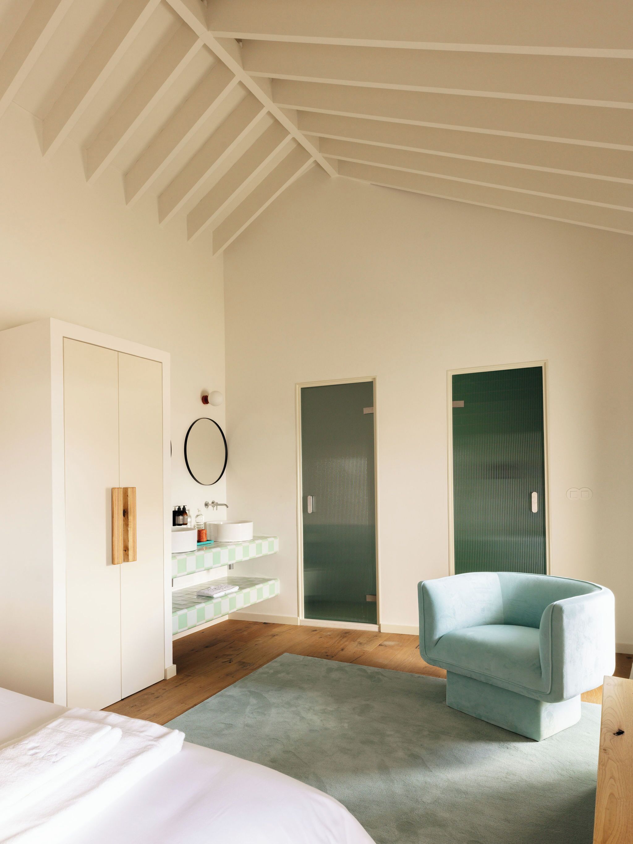 A bedroom with a bed, a green chair, a vanity with a mirror, and two doors.