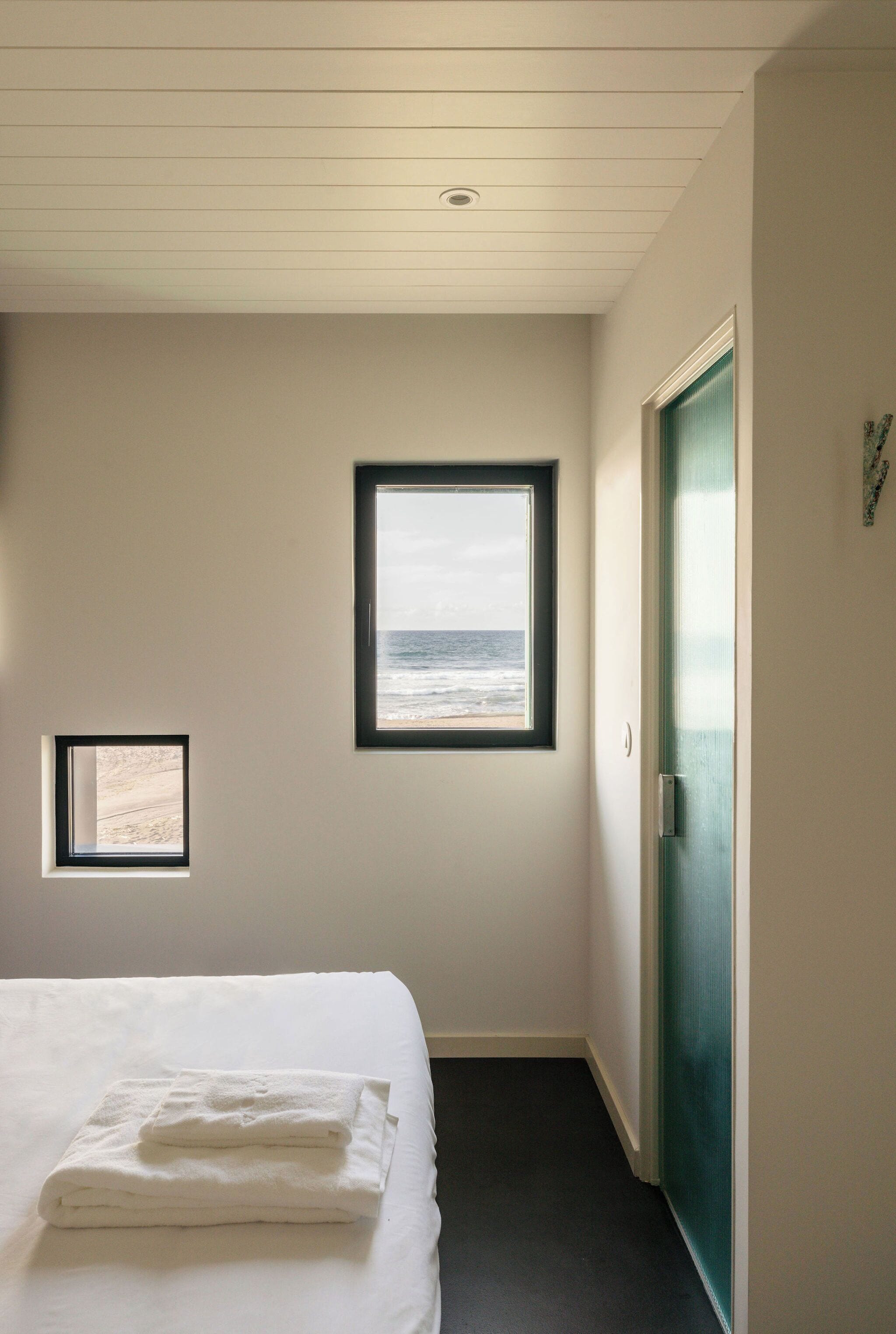 A hotel room with a bed, a small window with a beach view, and a door.