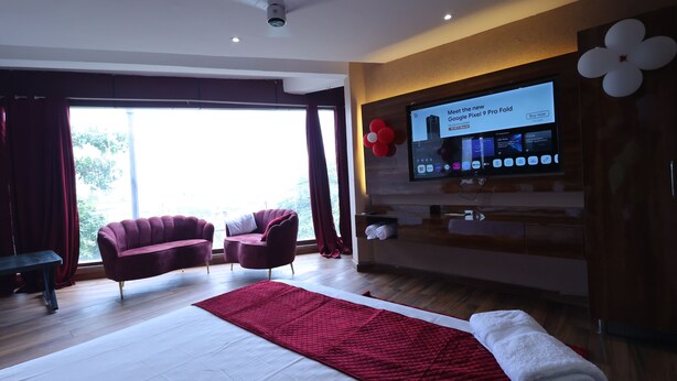 A modern hotel room with a large bed, a red quilted blanket, two armchairs, a flat-screen TV displaying a Google Pixel 9 Pro Fold advertisement, and a large window with curtains. - Hotel M2M  Narela (New Delhi)