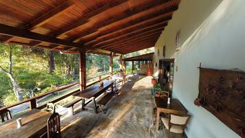 An outdoor covered patio with wooden benches and tables, surrounded by greenery and a view of the forest.