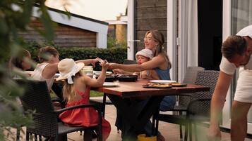 Una famiglia cena all'aperto a un tavolo con una griglia, circondata da sedie e un edificio moderno sullo sfondo.