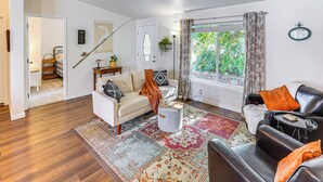 Ein Wohnzimmer mit einer Couch, einem Couchtisch und einem Teppich. Ein Fenster mit Vorhängen bietet Blick auf Grünfläche.