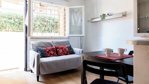 A compact living space with a sofa, a dining table with chairs, a window with a view of greenery, and a shelf with decorative items. - Montagnola Mini House (Rome)