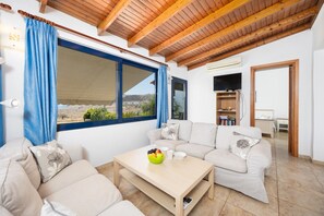 A modern living room with a sofa set, a coffee table with fruit, and a large window offering a view of the landscape.