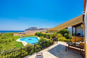 A pool with a view of the sea and a mountain, surrounded by greenery and a patio area with outdoor furniture.