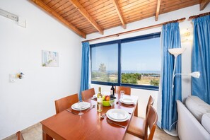 A dining area with a table set for four, a view of the sea through a large window, and a sofa in the corner.
