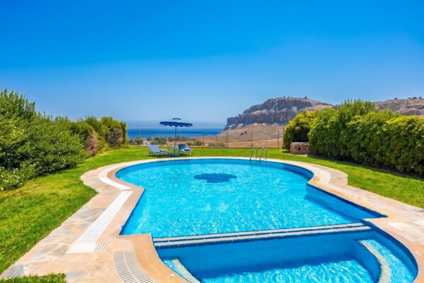 A swimming pool with a curved design, surrounded by well-maintained grass and hedges, with a view of the sea and mountains in the background.