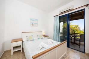 A bedroom with a bed, bedside tables, a nightstand, a window with curtains, and a view of a balcony with a table and chairs.