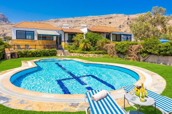 A swimming pool with a clear blue center, surrounded by a well-maintained lawn, and a view of a mountainous landscape in the background.