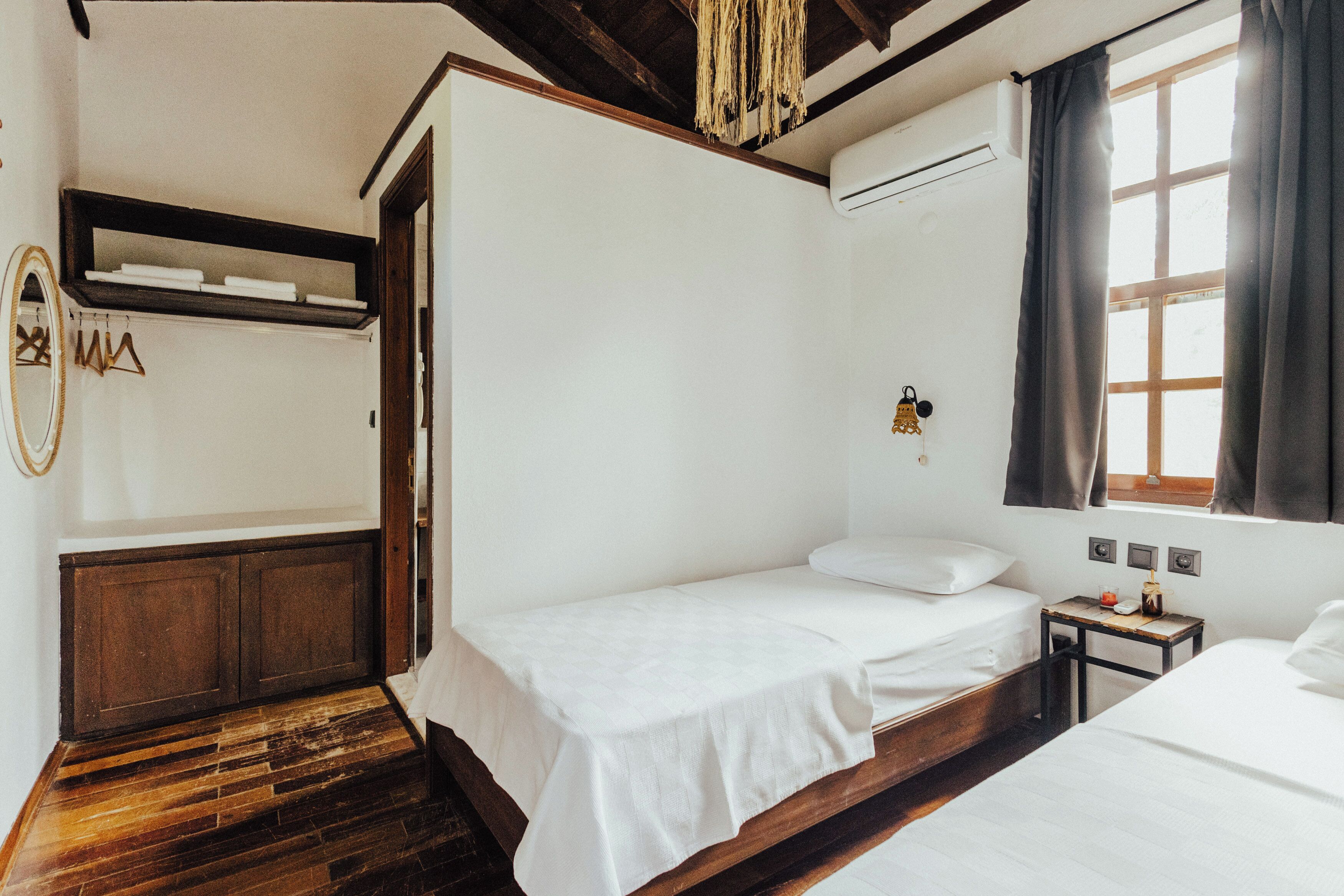 A room with two single beds, a wooden floor, a small table with a lamp, and a window with curtains.