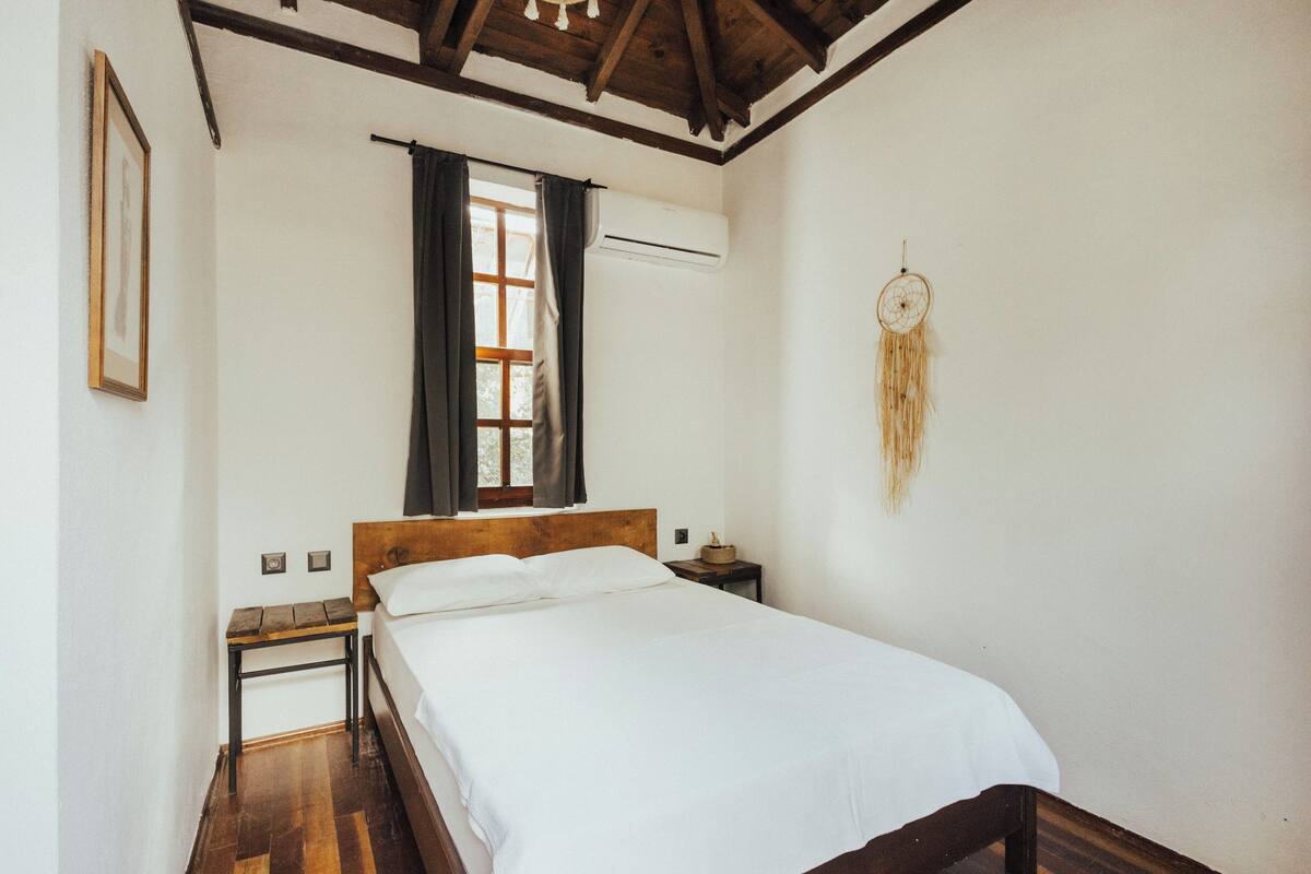 Ein Schlafzimmer mit einem hölzernen Bett, einem Nachttisch, einem Fenster mit Vorhängen und einem Traumfänger an der Wand.