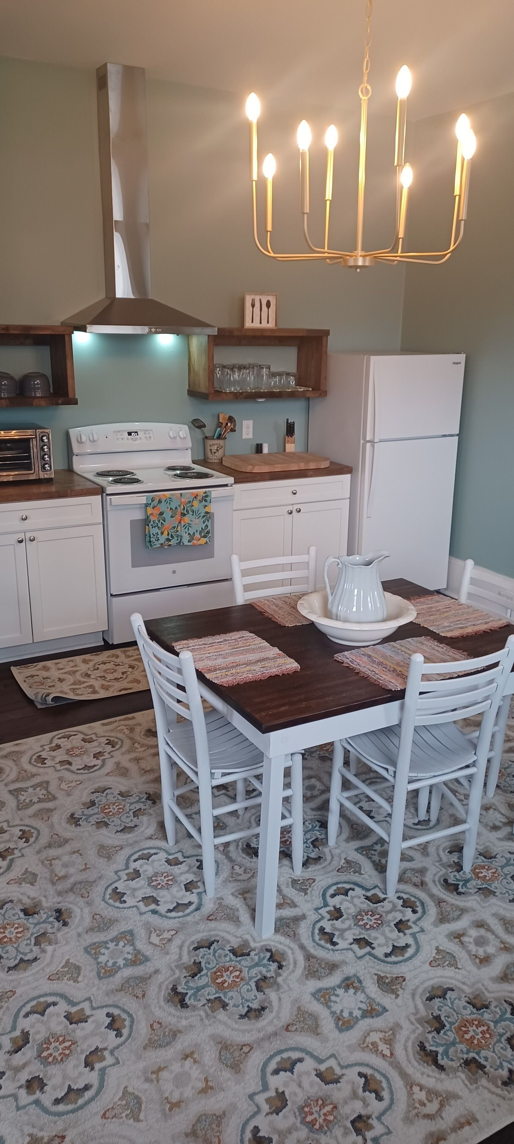 A dining area with a wooden table, white chairs, a chandelier, and a patterned rug.