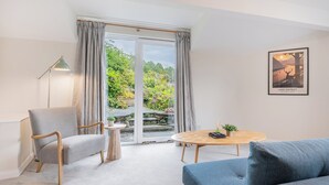 Una sala moderna con un sillón gris, un sofá azul, una mesa de centro de madera y vistas a un jardín a través de un ventanal corredero.