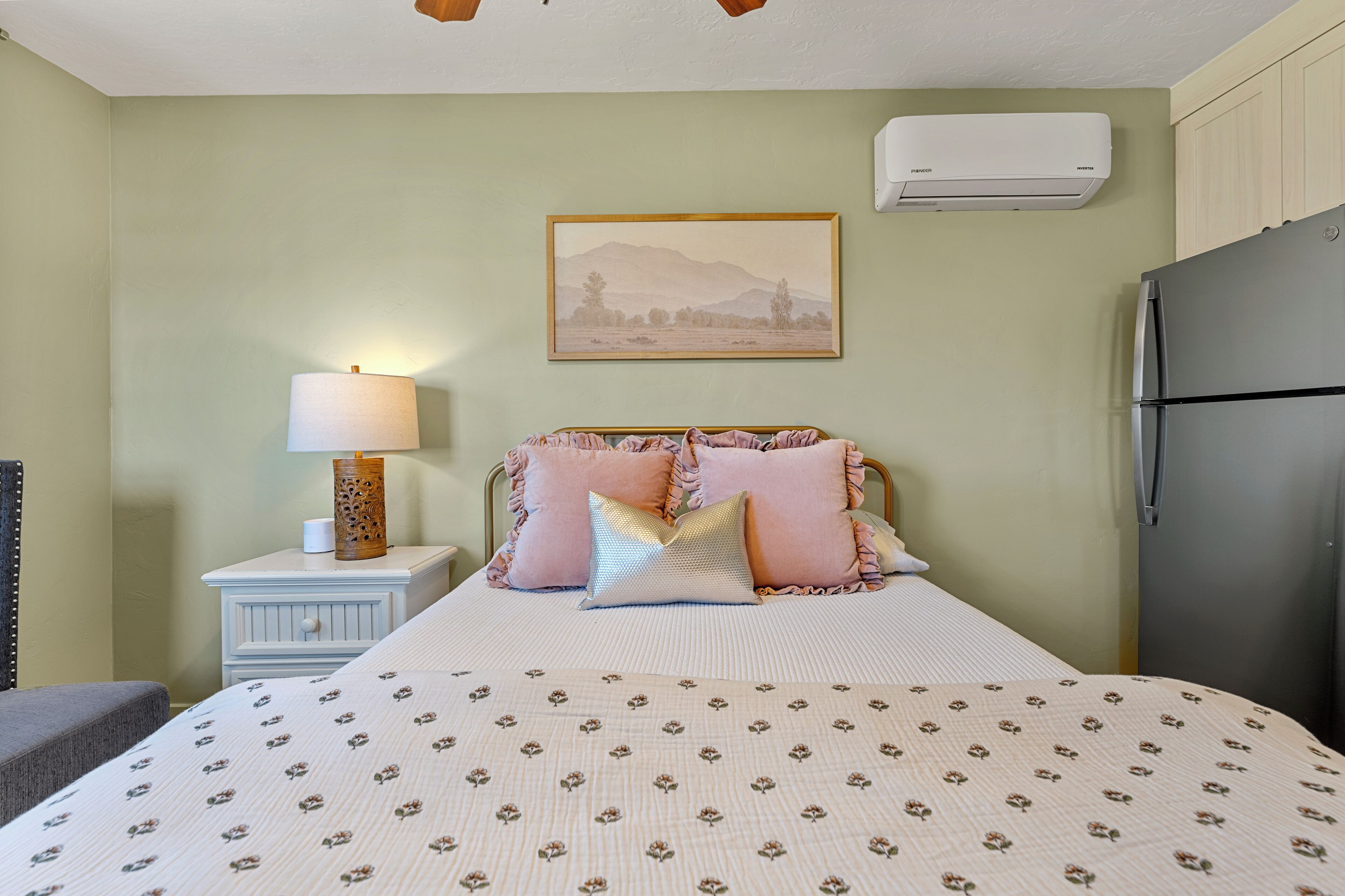 A bedroom with a bed, bedside table with a lamp, a refrigerator, and a wall-mounted air conditioner.
