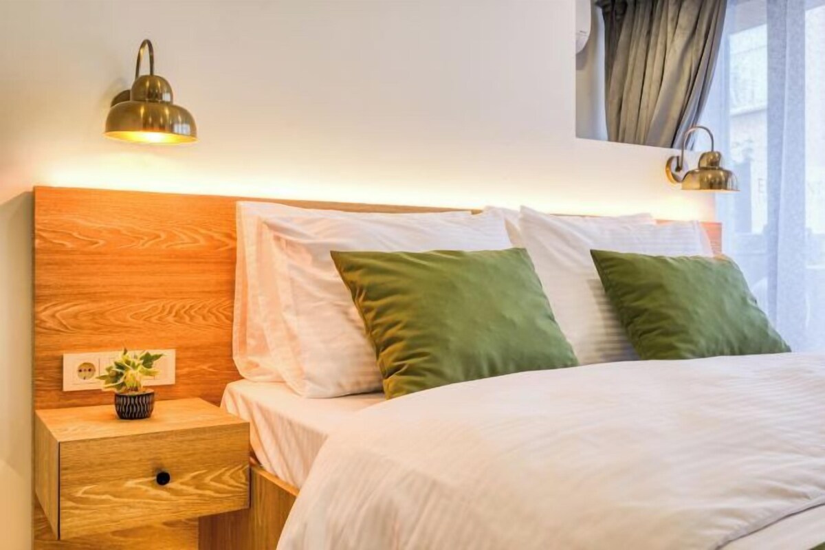 A neatly made bed with white and green pillows, a wooden headboard, and a bedside table with a small plant.