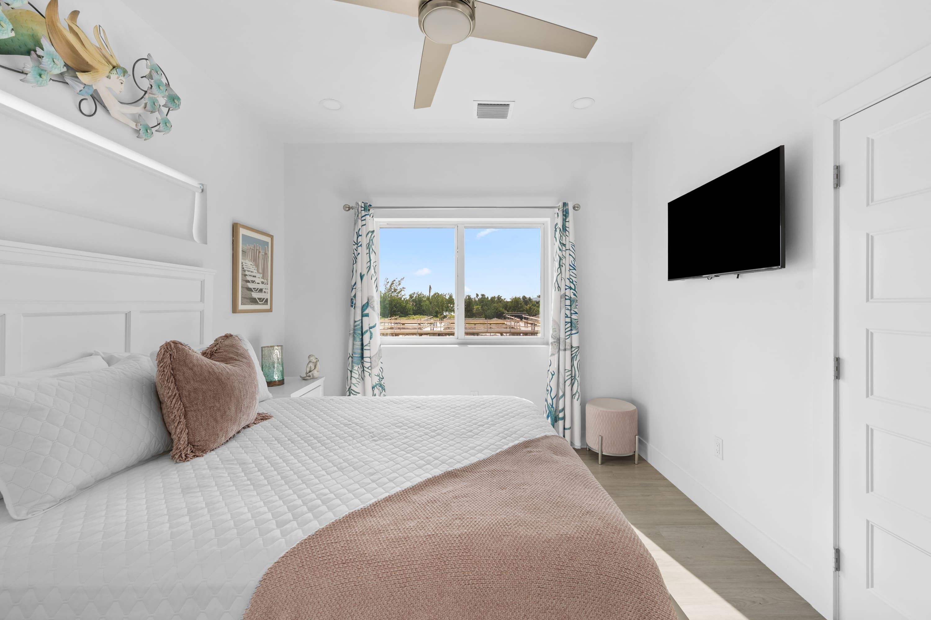 A bedroom with a bed, a ceiling fan, a TV, a window with curtains, and a door.