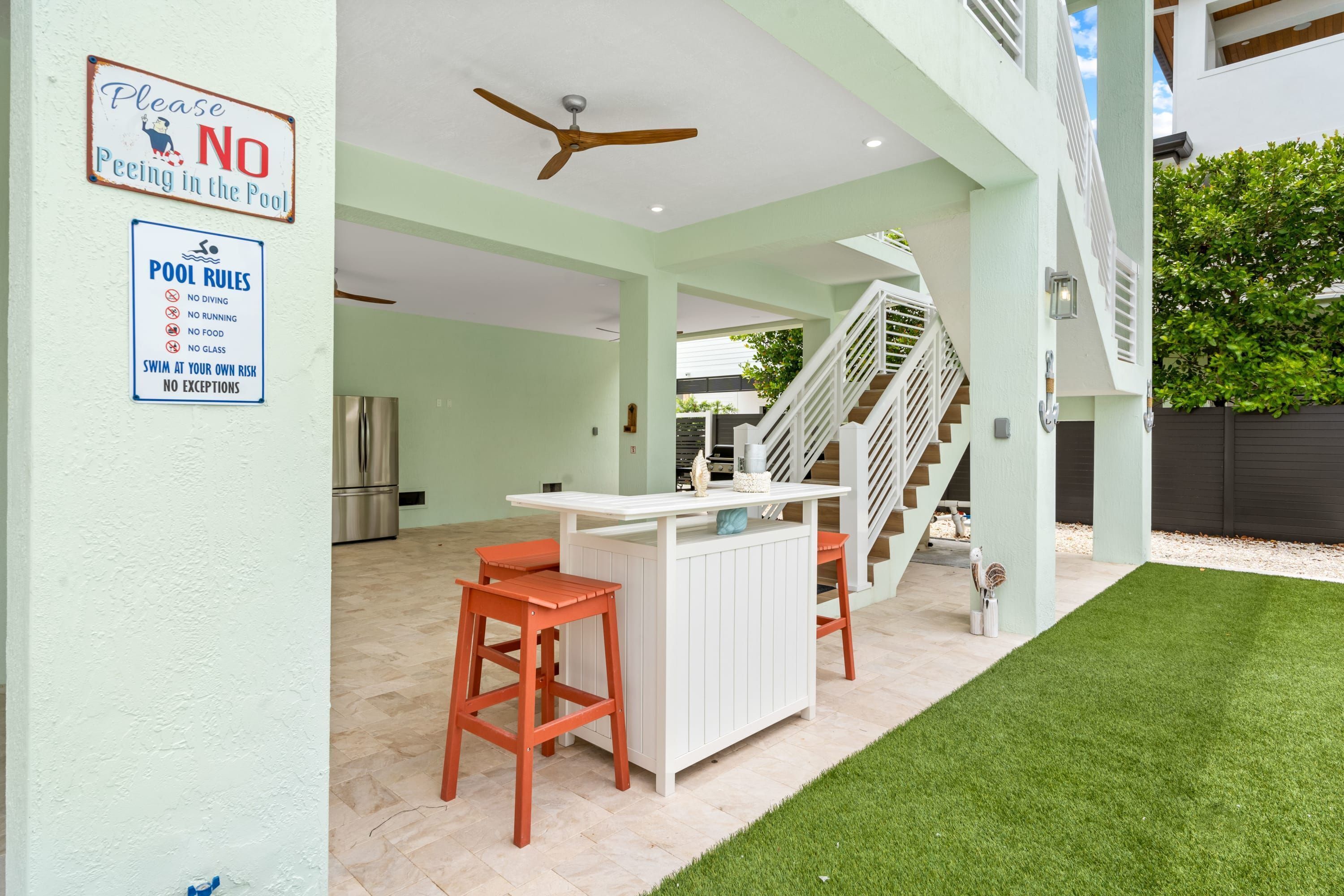 Zona de piscina con un cartel de prohibición de orinar, normas de la piscina, una zona de bar blanca con taburetes, un ventilador de techo y una escalera que conduce a un nivel superior.