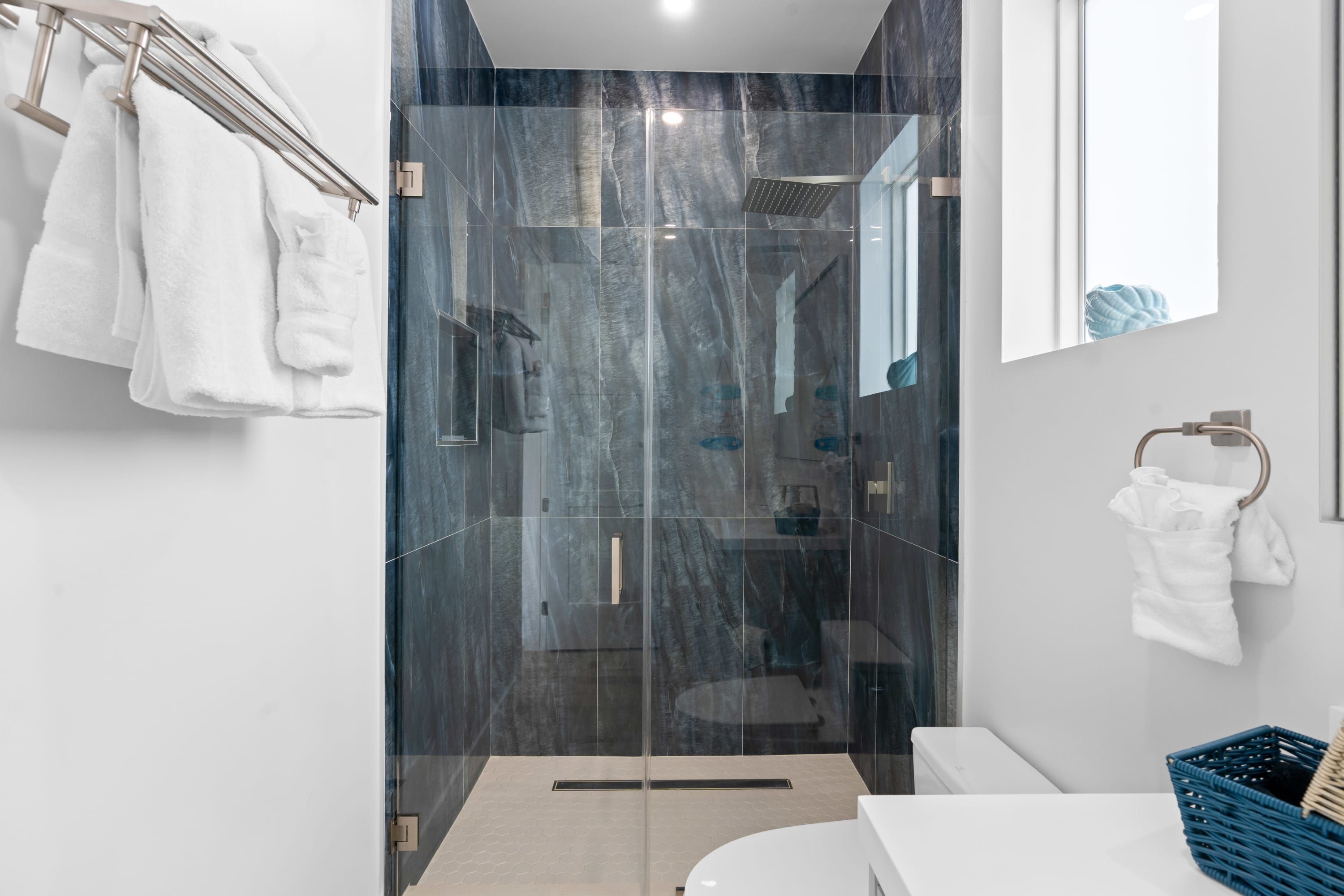 Un baño moderno con ducha envuelta en vidrio, sanitarios blancos y una cesta de mimbre azul.
