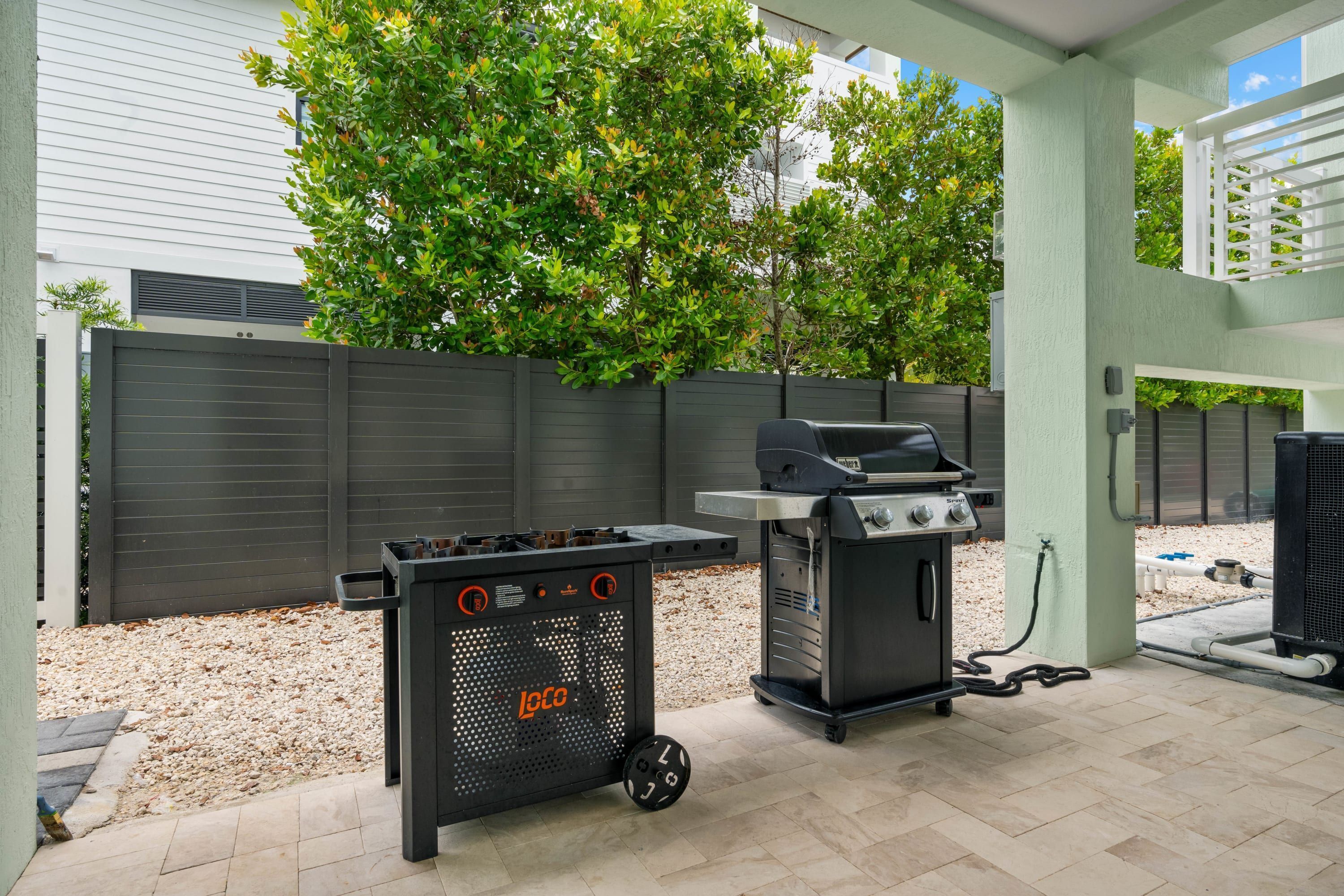 Un espace patio équipé d’un barbecue et d’un chariot de cuisson mobile, le tout entouré d’une clôture et d’arbres.