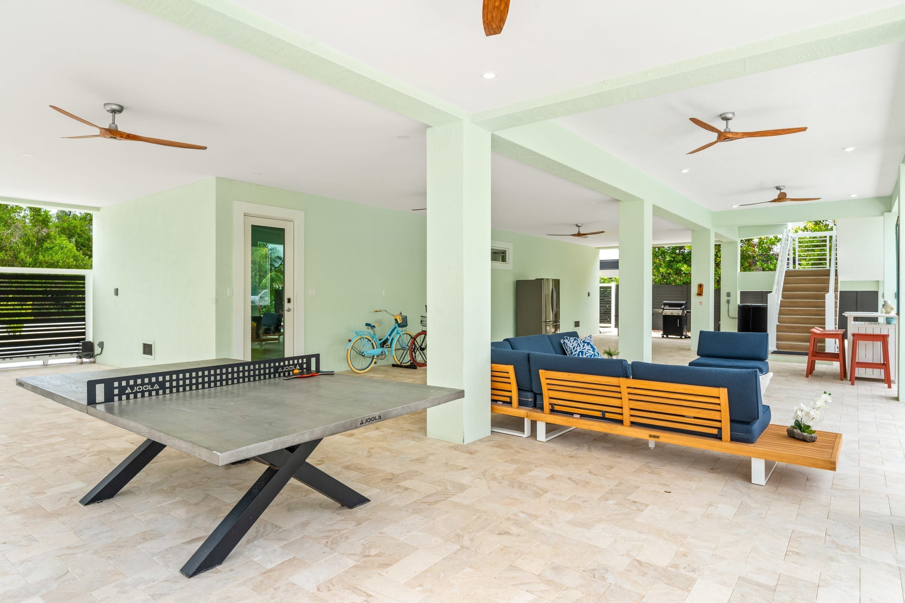 A spacious living area with a ping pong table, a sofa, and a wooden bench. There are ceiling fans, a bicycle, and a kitchen area visible.