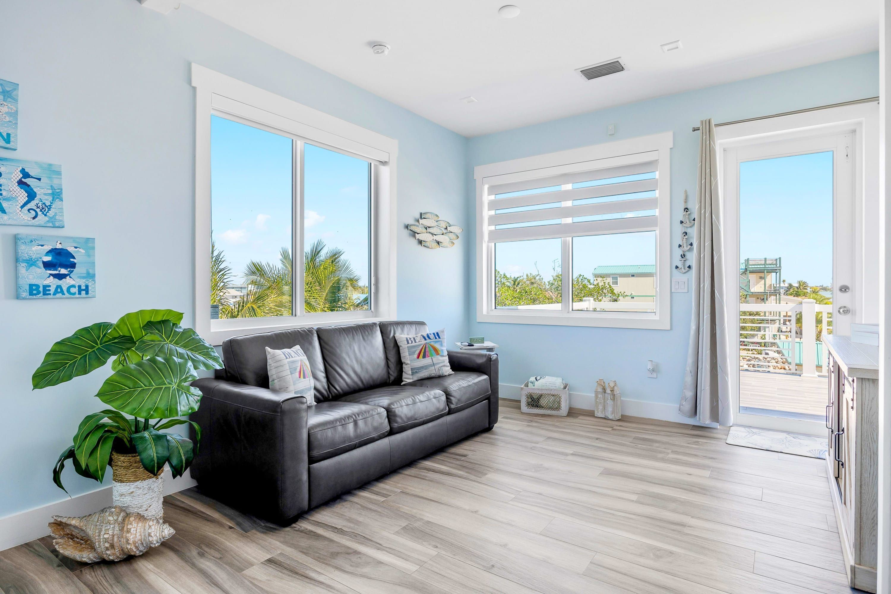A modern living room with a black leather sofa, large windows, a potted plant, and a view of the beach.