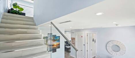 A modern interior with a staircase, a glass railing, a wooden floor, a glass console table, and a round mirror on the wall.