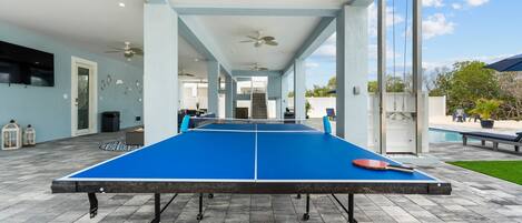 A ping pong table with a paddle, set up on a patio with a pool in the background.