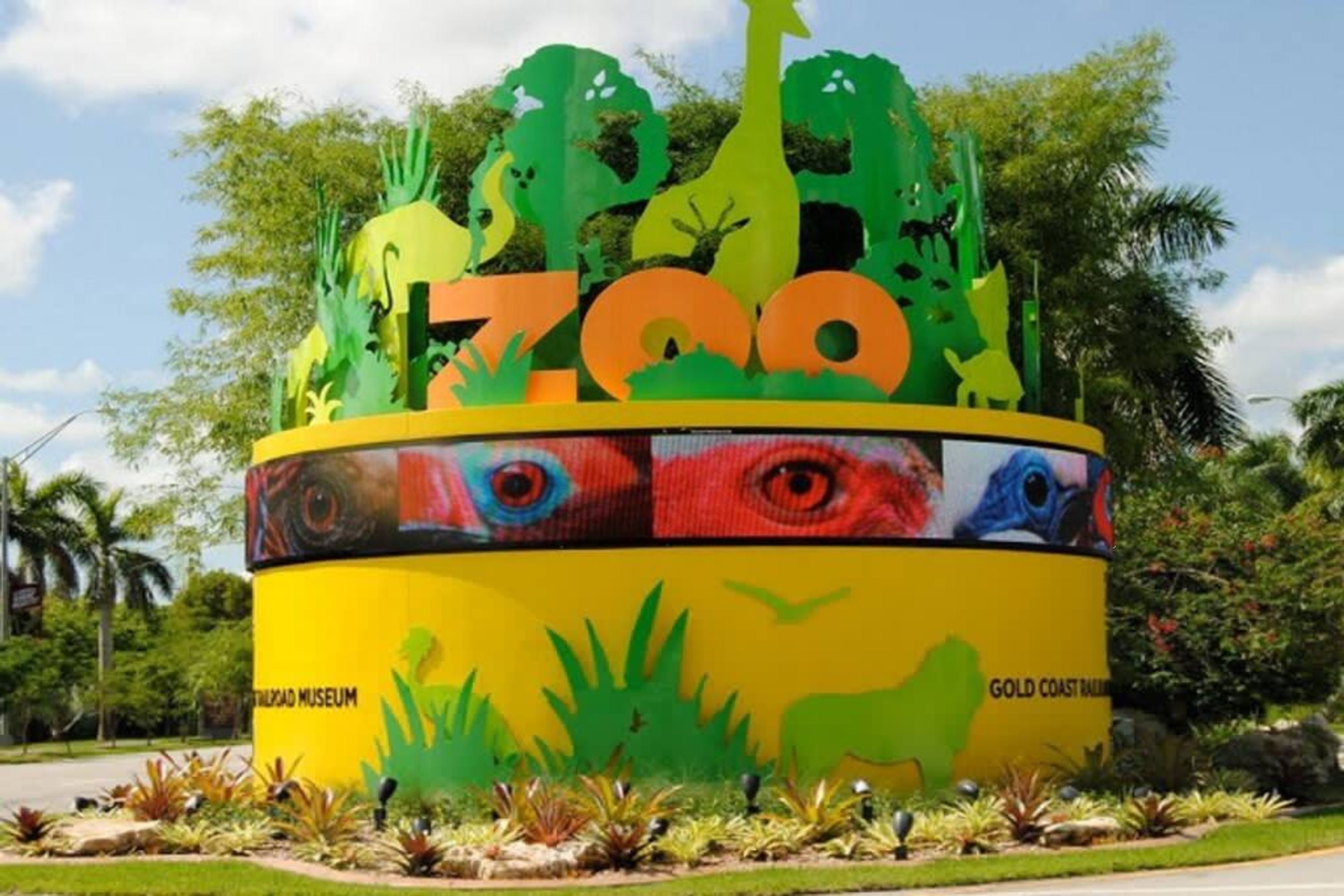 Un letrero circular y colorido con la inscripción 'ZOO' y 'Gold Coast Zoo', rodeado de plantas y árboles tropicales.
