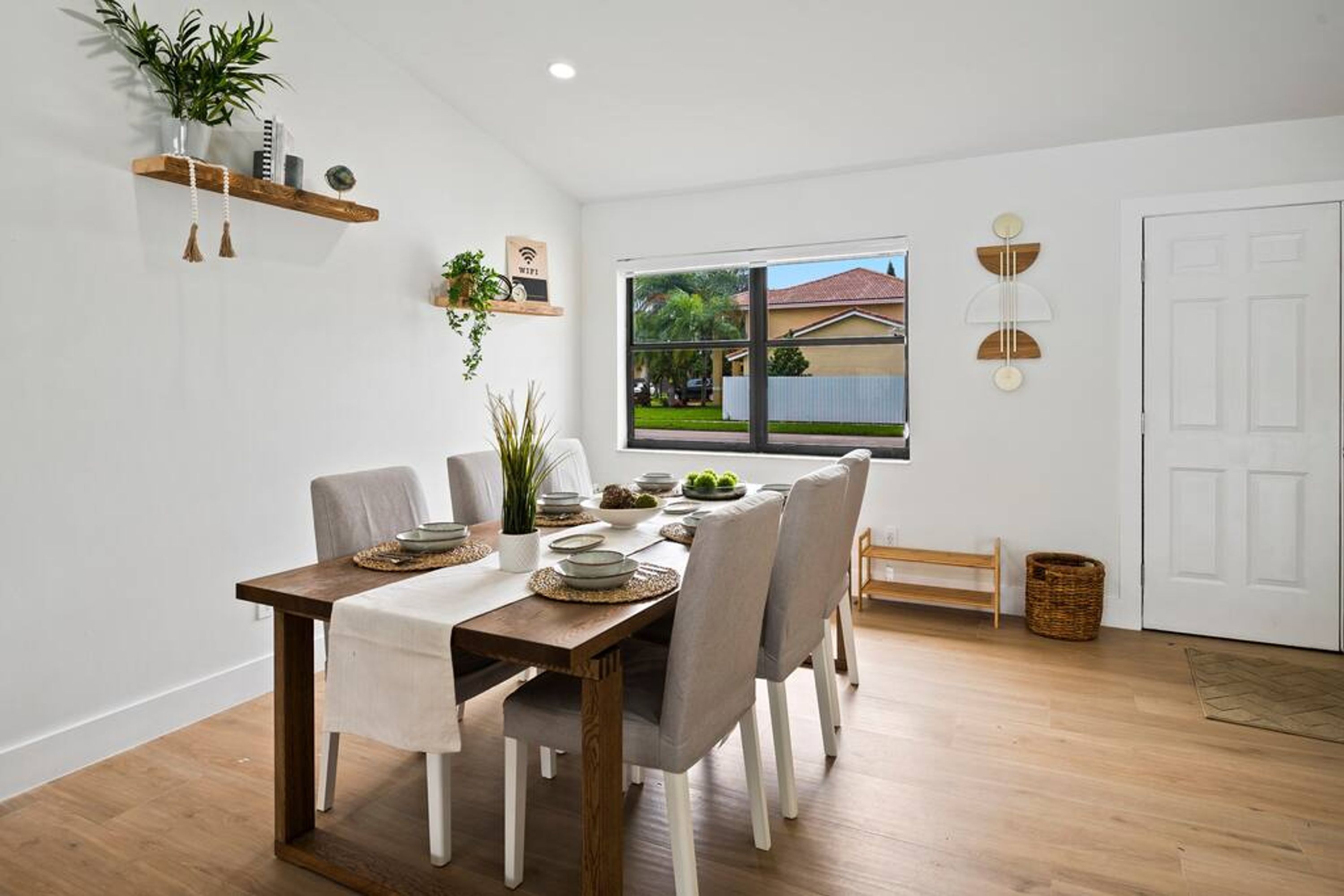 Un coin repas avec une table en bois, des chaises à revêtement gris et une vue sur une maison, visible à travers une grande fenêtre.