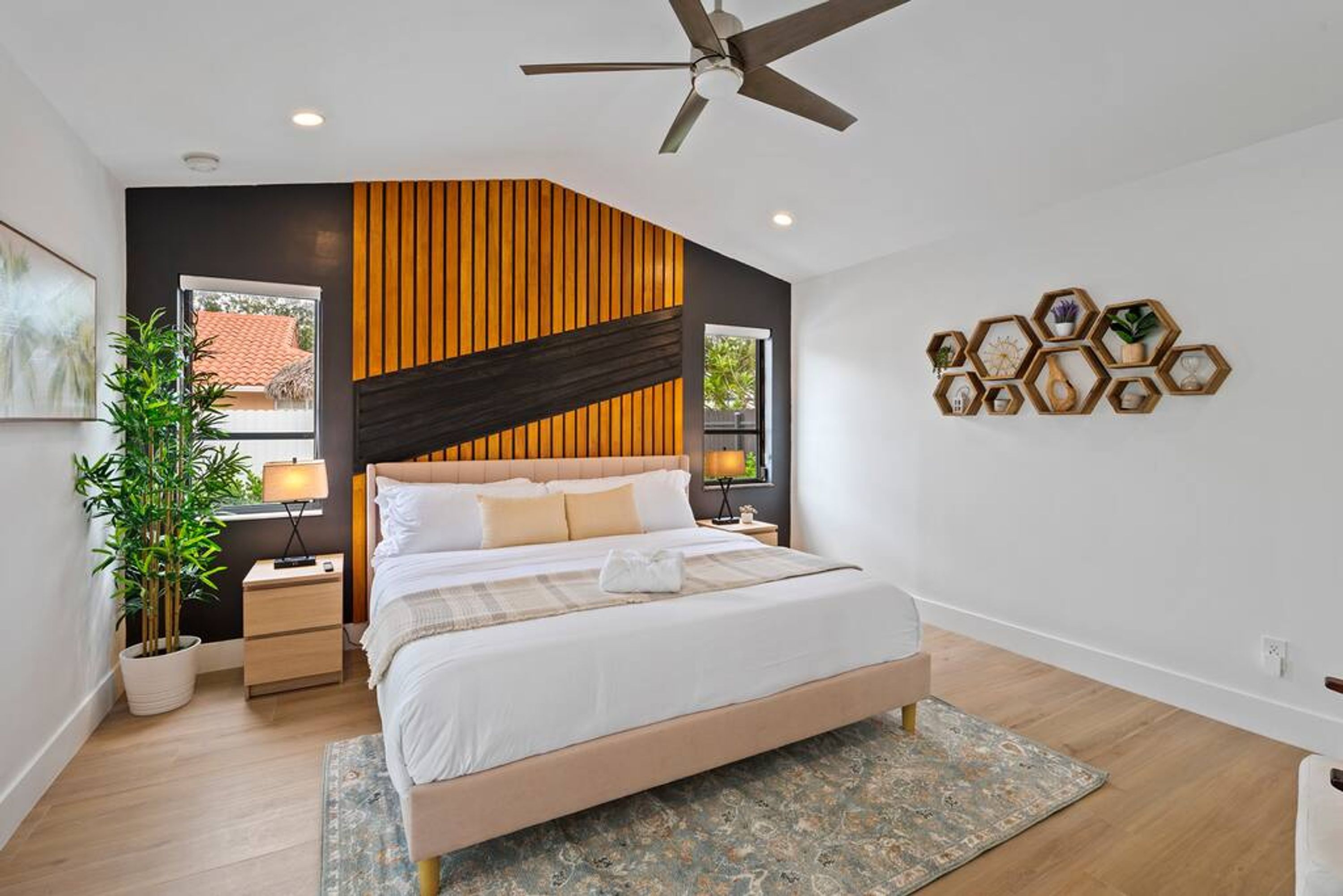 Une chambre à coucher moderne, avec un lit, des tables de chevet, un ventilateur de plafond et une étagère hexagonale fixée au mur, agrémentée d’objets décoratifs.