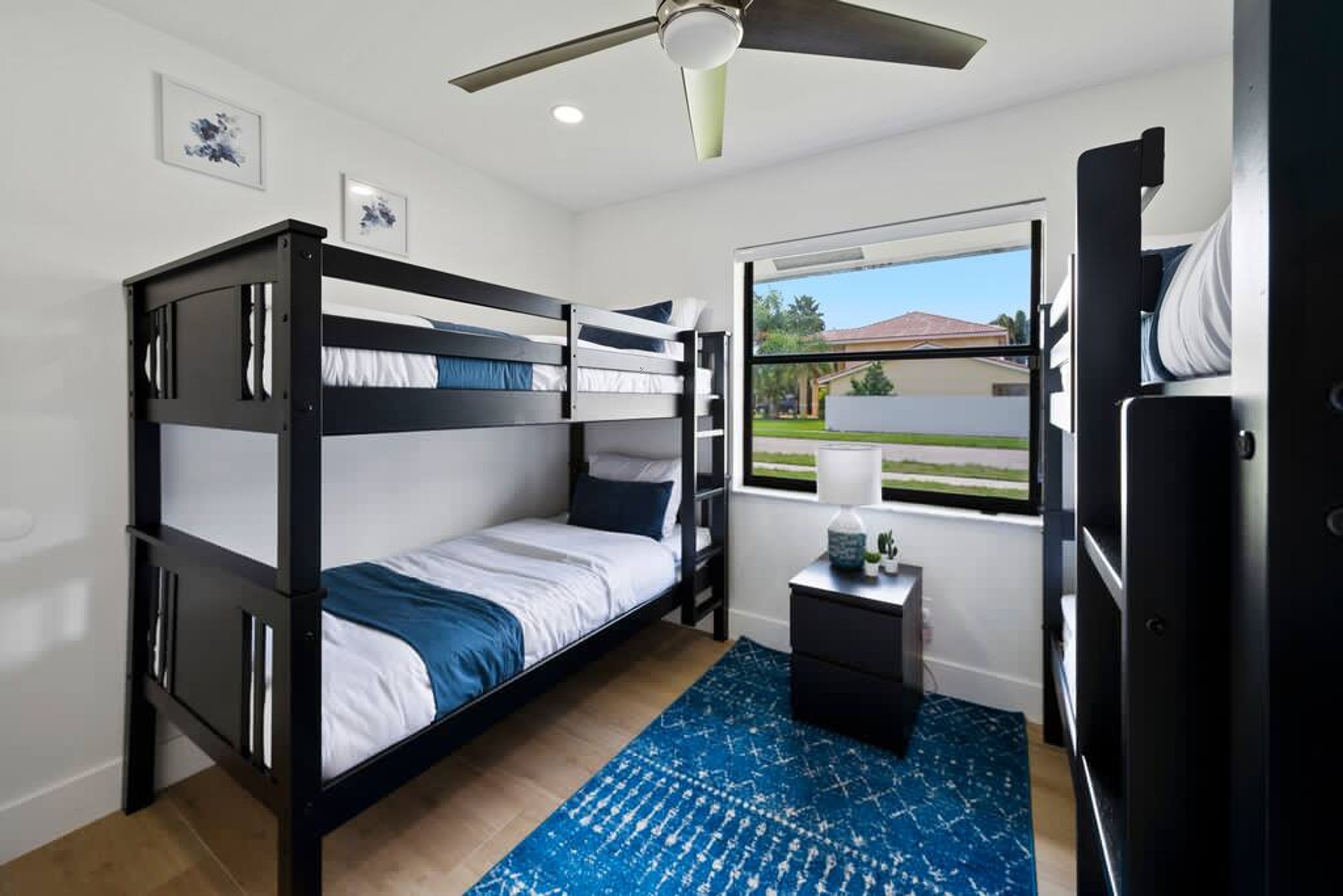 Habitación con literas, ventilador de techo, una mesita con lámpara y ventana con vistas a una casa y árboles.