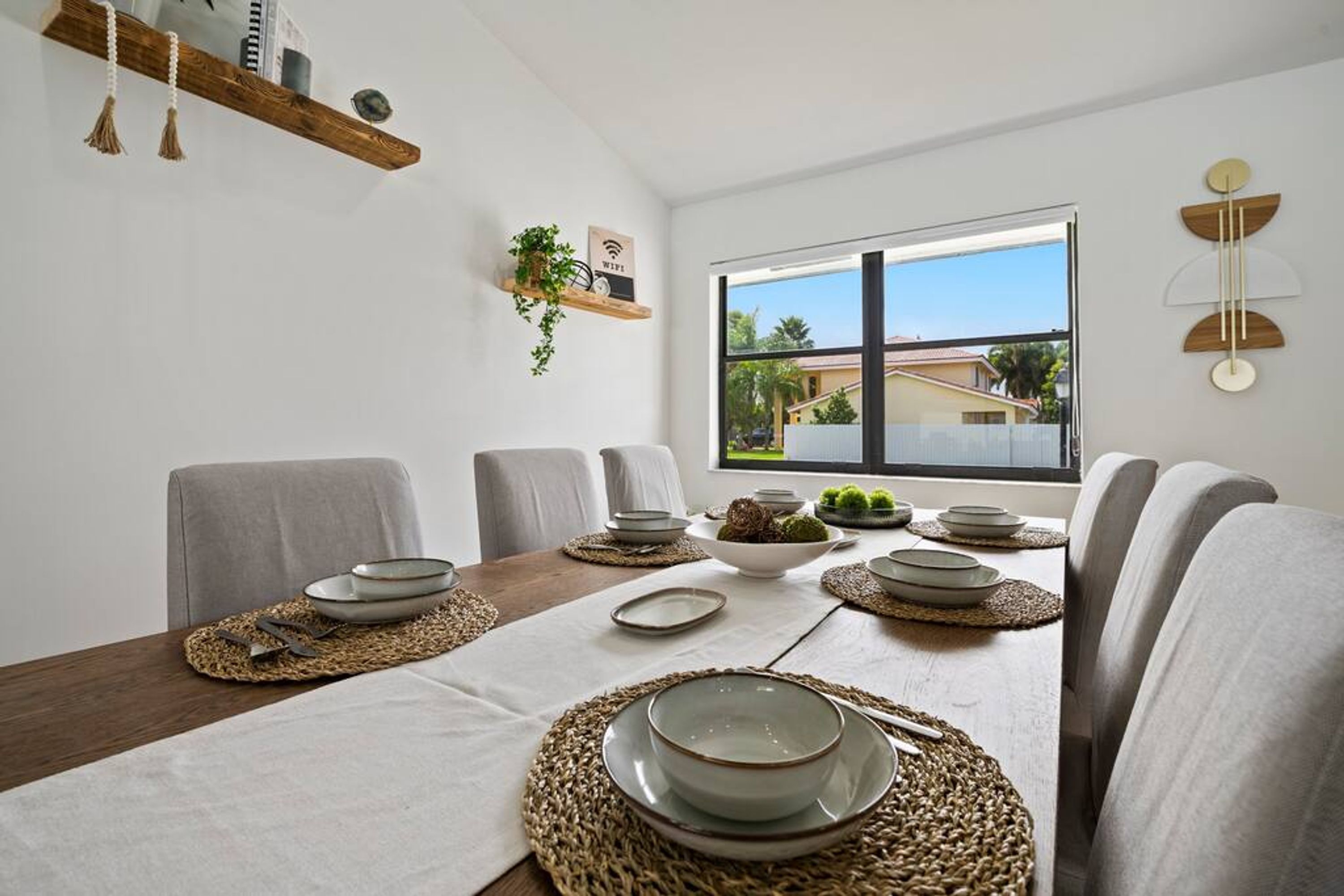 Comedor con una mesa de madera dispuesta para seis comensales, sillas blancas y un ventanal con vista a edificios.