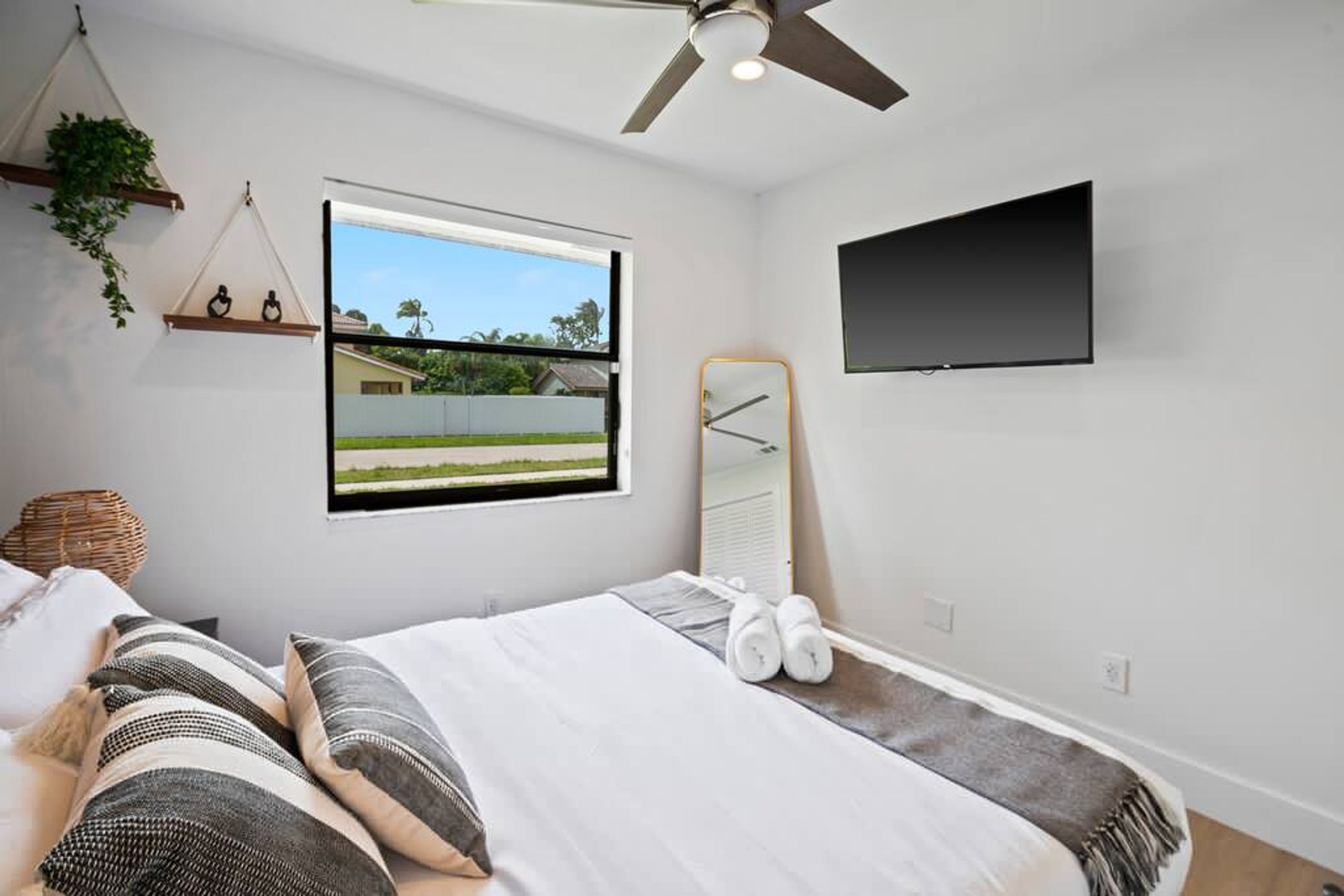 Une chambre avec un lit, un ventilateur de plafond, un téléviseur fixé au mur, une fenêtre donnant sur une maison et de la verdure, et une plante suspendue.