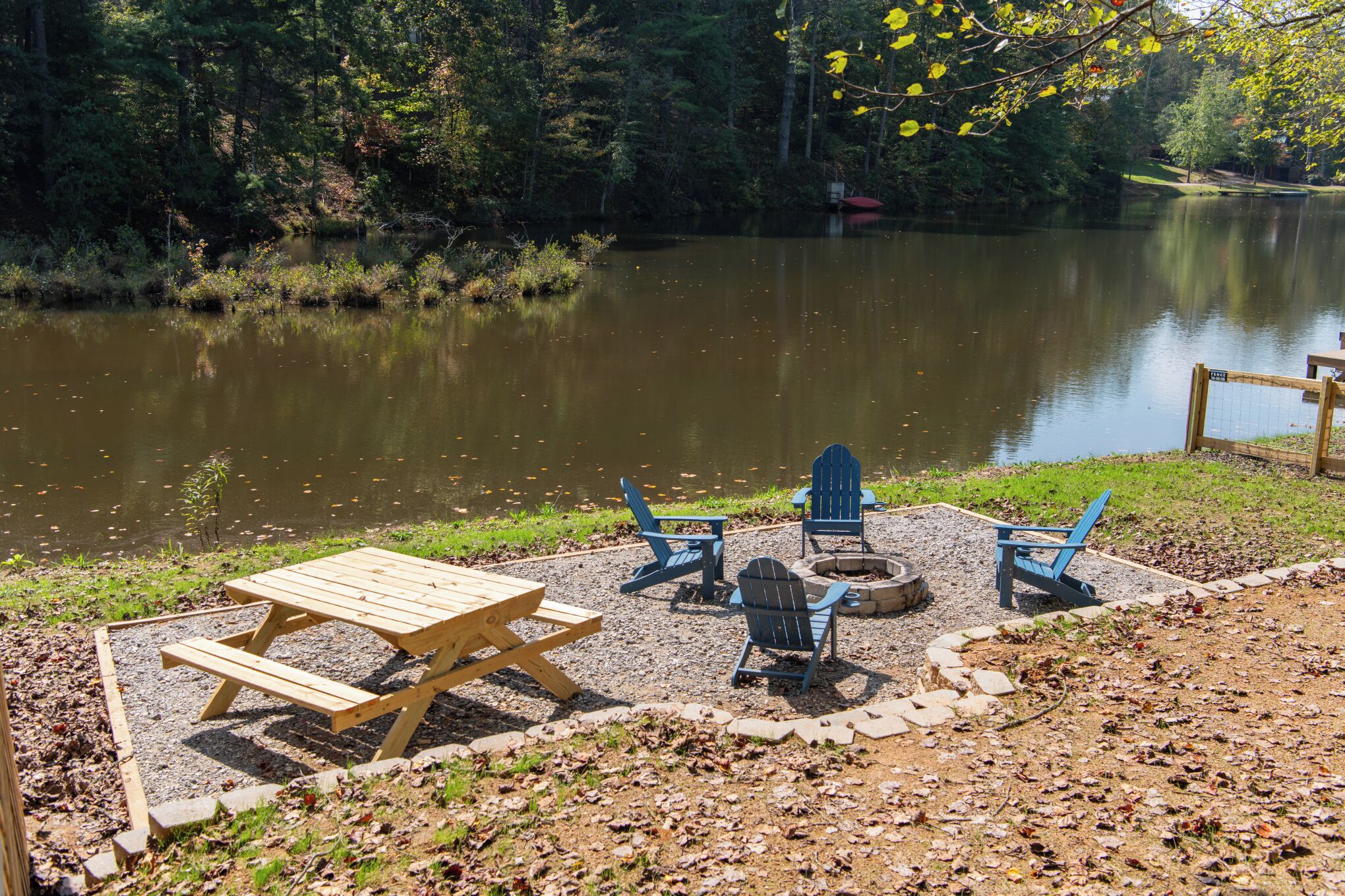 A lakeside picnic area with a wooden table and chairs, a fire pit, and a stone border.