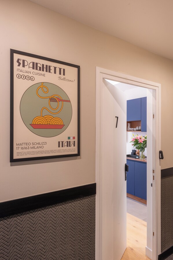A hallway with a framed poster of spaghetti, a door labeled '7', and a glimpse of a kitchen area.