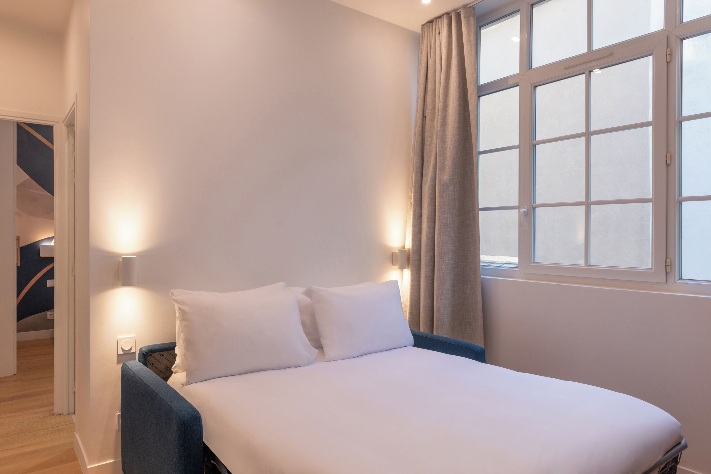 Een slaapkamer met een bed, twee bedlampen, een raam met gordijnen en een trap op de achtergrond.