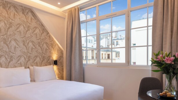 Habitación de hotel con una cama grande, una mesita con una silla, un jarrón con flores y una ventana con vistas a edificios.