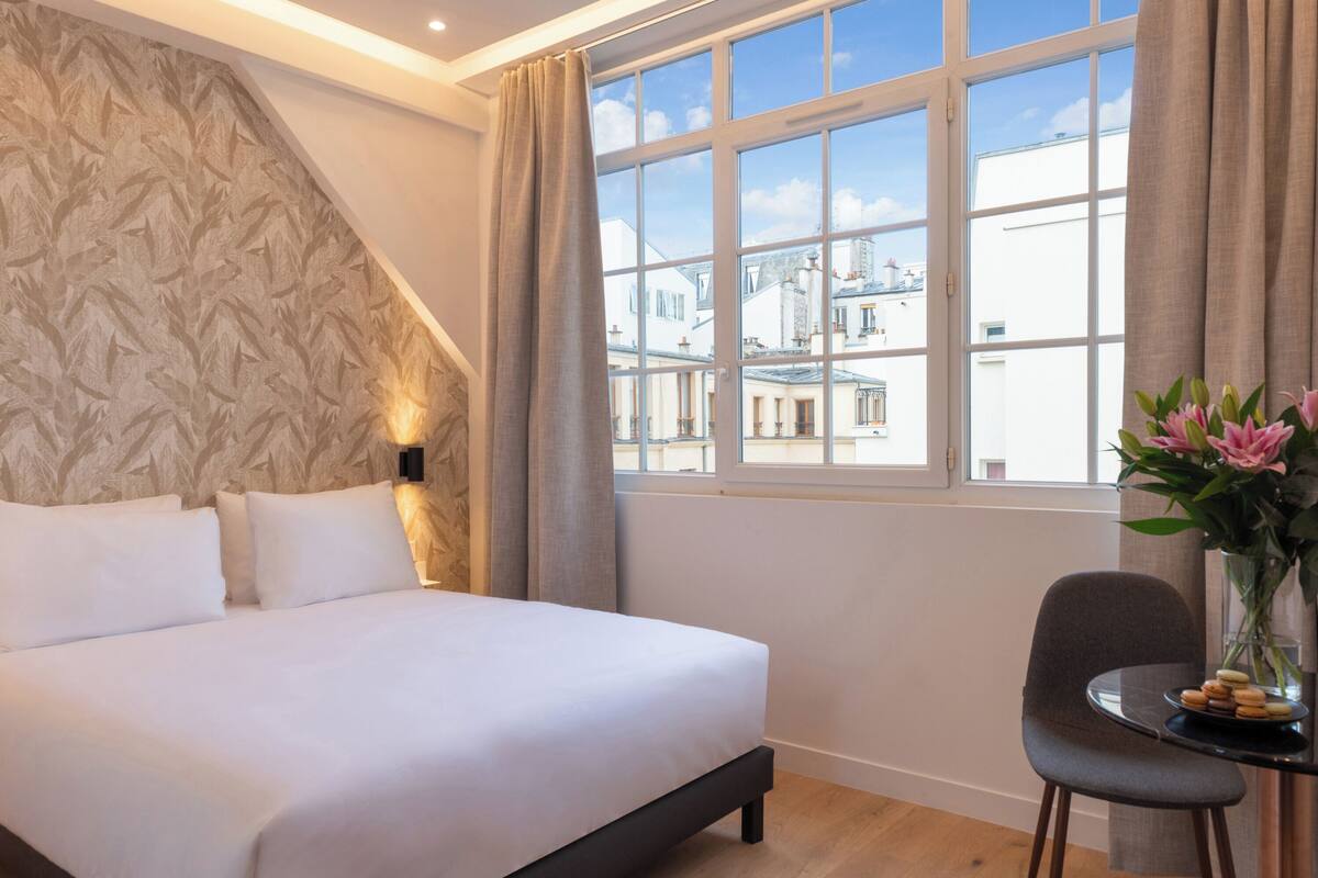 A hotel room with a large bed, a small table with a chair, a vase with flowers, and a window with a view of buildings.