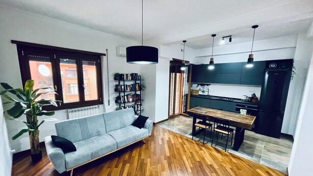 A modern living room with a sofa, wooden flooring, a bookshelf, and a kitchen area with a refrigerator and cabinets. - New apartment in Rome Trastevere w/ 2 beds, living, excellent transport (Roma)