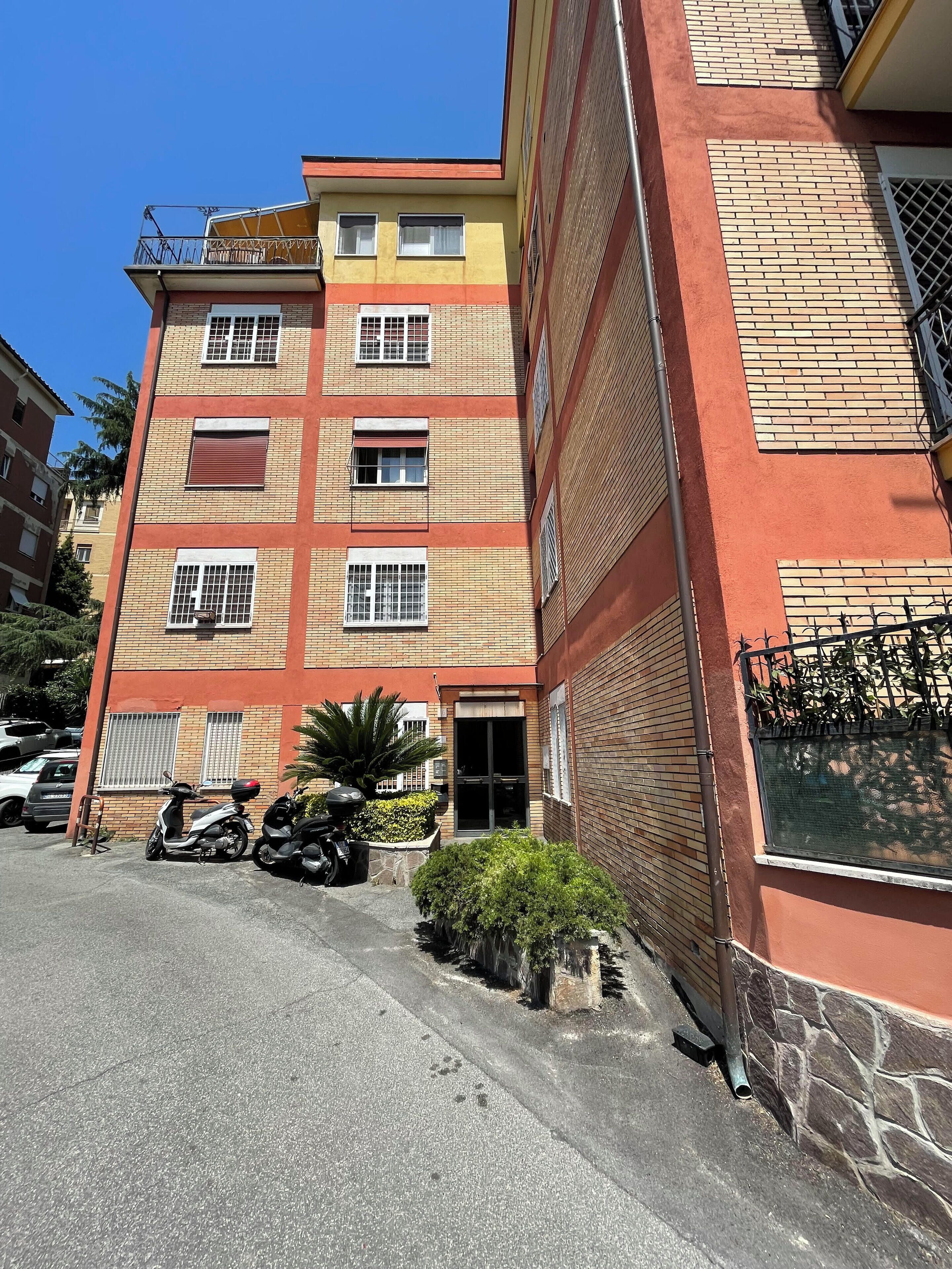 Un edificio a più piani con facciata in mattoni, un ingresso con cancello e un'area parcheggio con scooter.