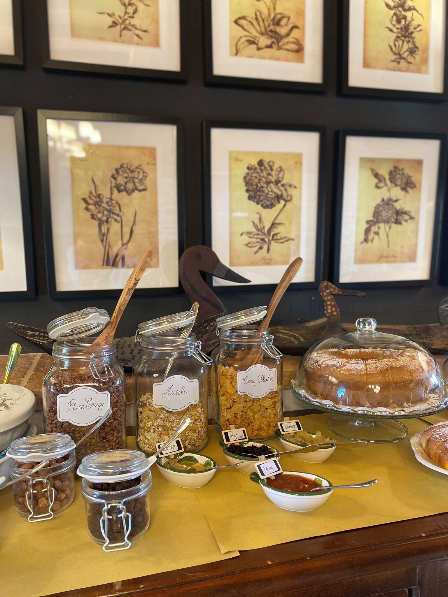 A display of food items with labels, a cake on a stand, and framed botanical illustrations in the background.