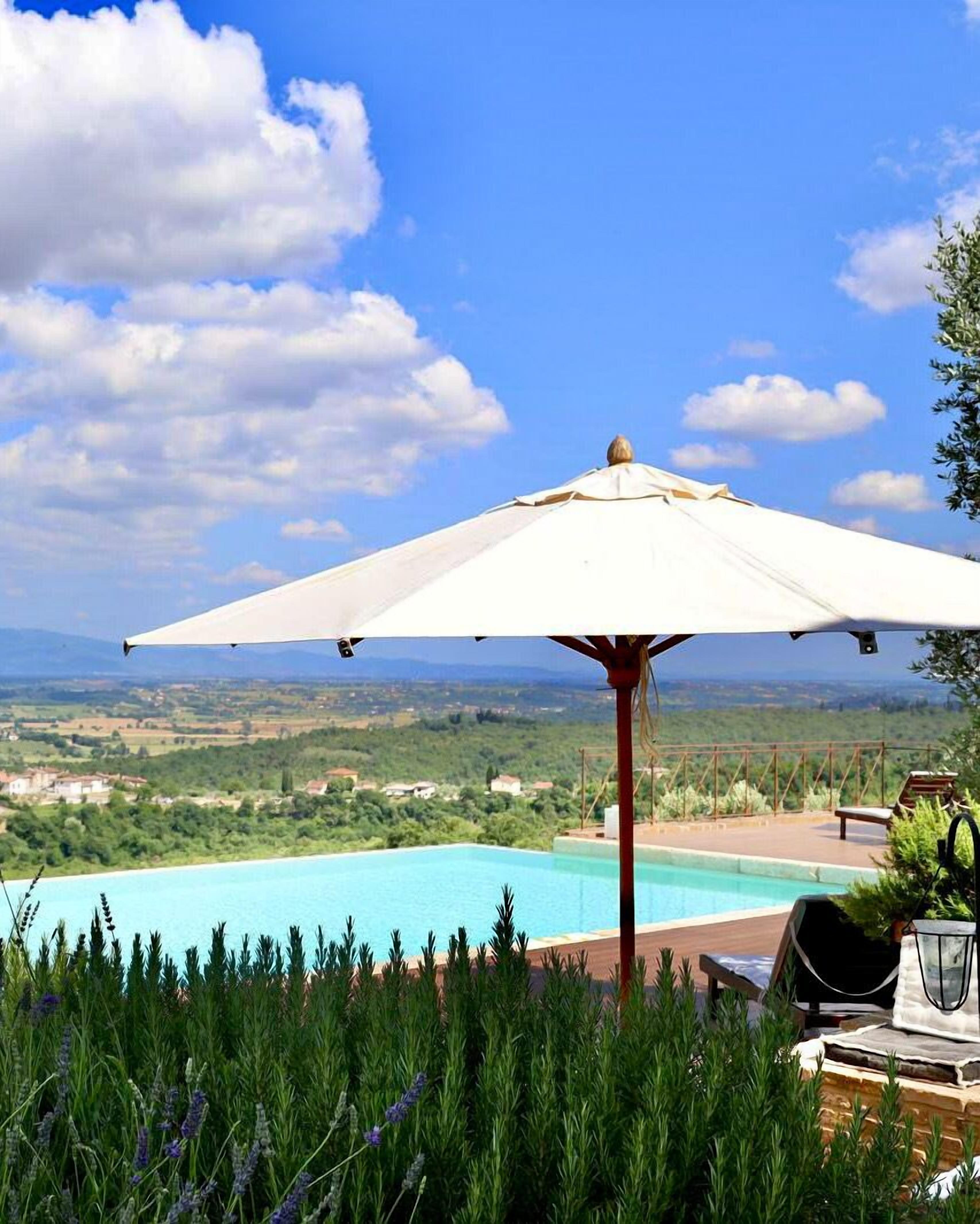 A poolside area with a white umbrella, a view of a pool, and a landscape in the background.