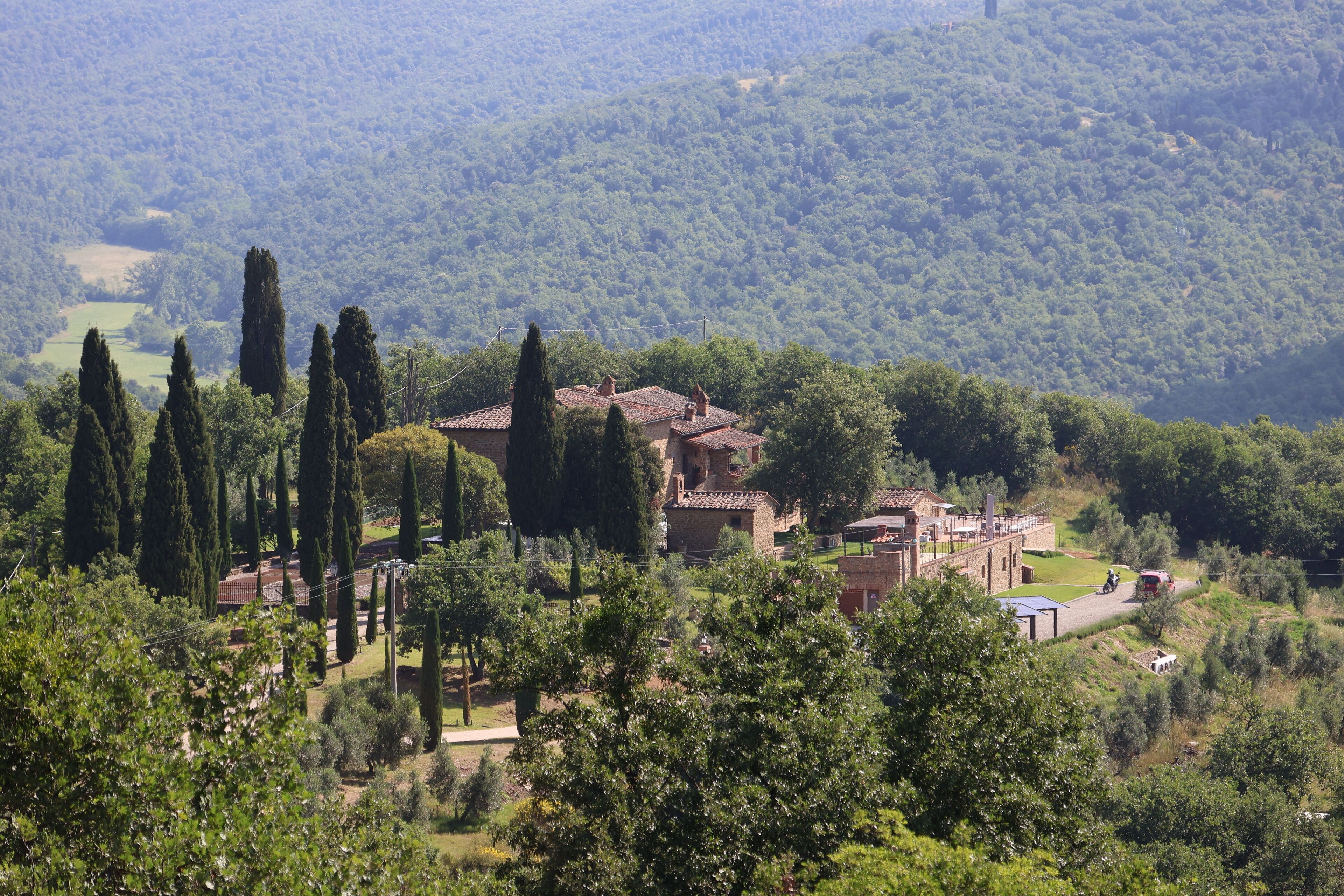 A rural villa with a courtyard, surrounded by tall cypress trees and a hilly landscape.