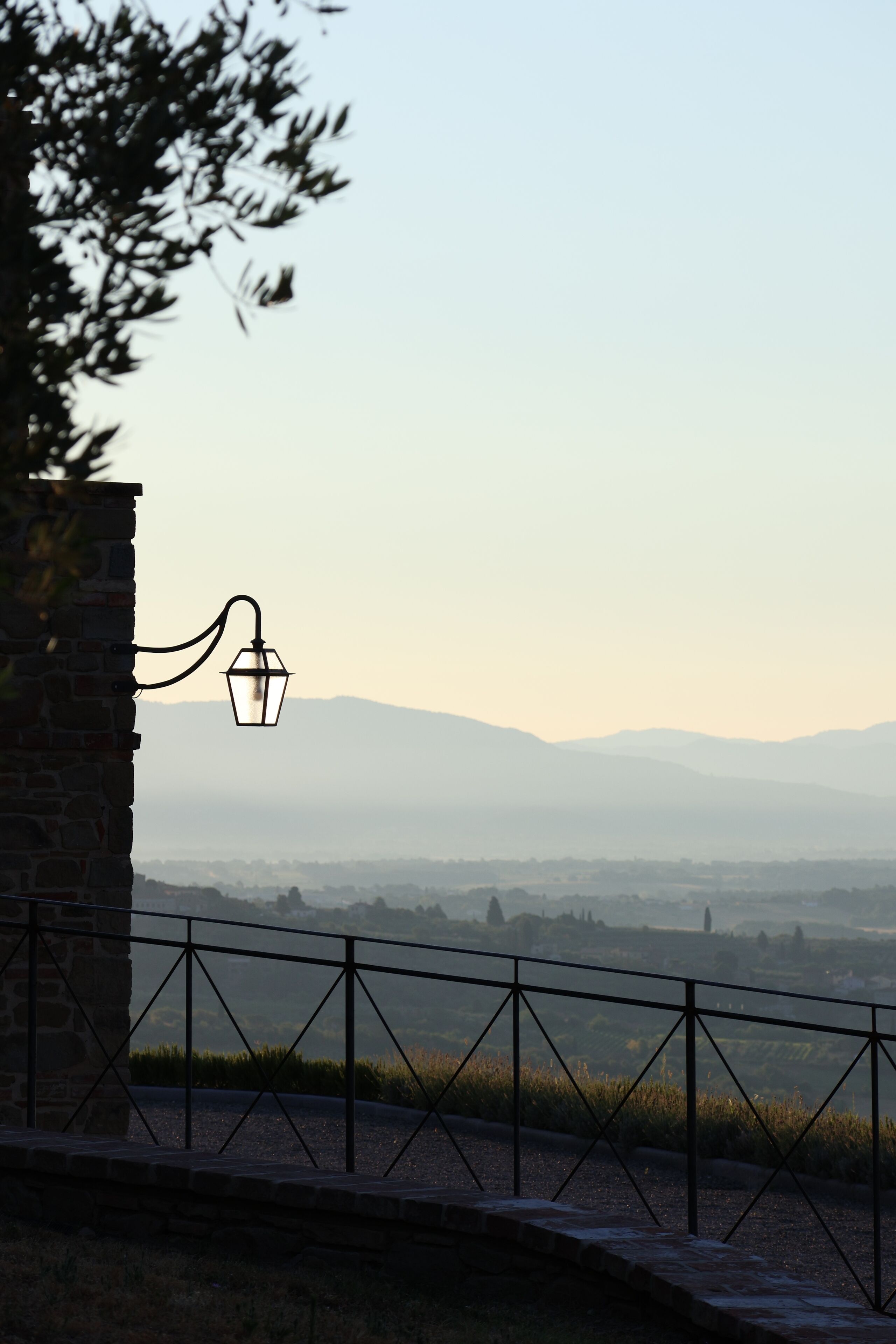 A stone wall with a metal railing and a hanging light fixture overlooking a landscape with distant hills.