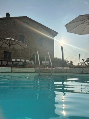 A swimming pool with clear water, a stone building in the background, and two umbrellas. - Podere Cavalbianco (Monte San Savino)