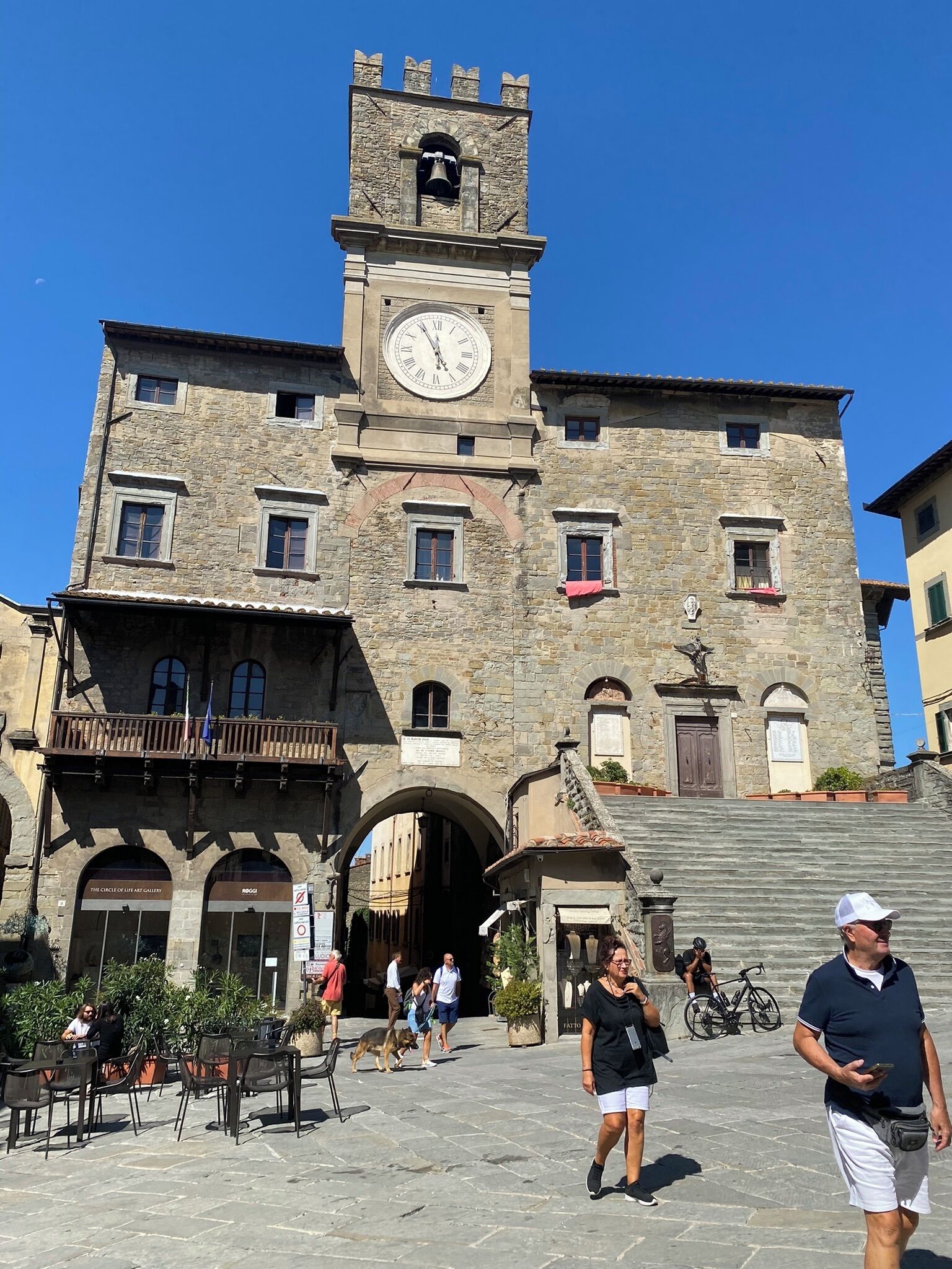 Un edificio storico in pietra con una torre dell'orologio, un arco d'ingresso e posti a sedere all'aperto.